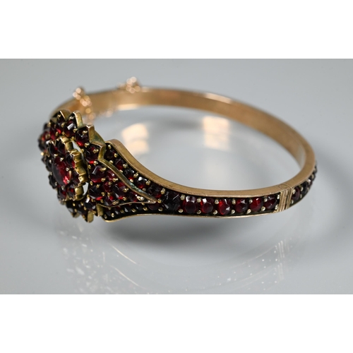 243 - A Victorian garnet-set bangle, the oval half-hinged yellow metal bangle with central flower motif, c... 