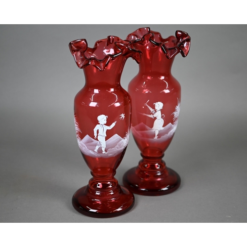 556 - A pair of cranberry glass vases with crenellated rims and waisted stems, painted with children playi... 