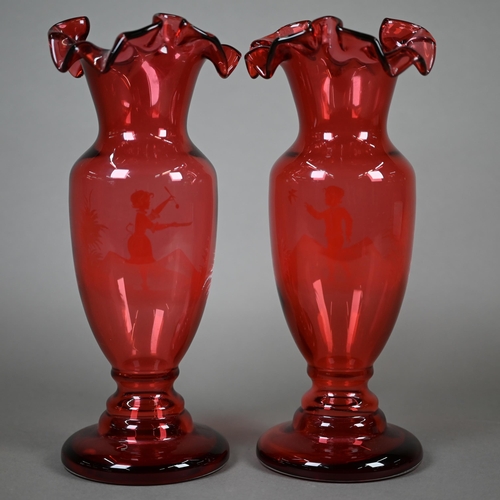 556 - A pair of cranberry glass vases with crenellated rims and waisted stems, painted with children playi... 