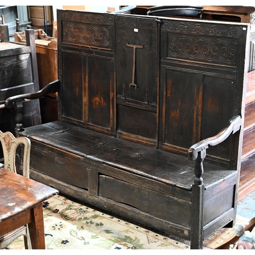 A 17th century oak box settle, the crest rail with lunette carved decoration over further carved panels, unusually centred by a hinged panel folding to form a table top, with iron fork form prop, the shaped arms over a twin hinged plank seat enclosing a storage well, raised on moulded stiles, 194 cm wide x 64 cm x 150 cm h (the seat 47 cm h)