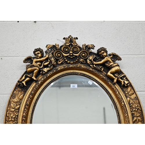 223 - A modern oval wall mirror in decorative gilt frame with foliate designs surmounted by cherubs, 98 cm... 