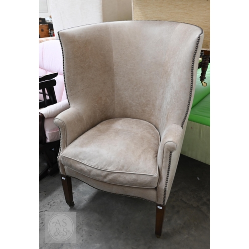 An Andrew Martin Georgian-style rounded wingback library armchair upholstered in studded buff suede, raised on stained square hardwood legs with brass casters, originally supplied by Hepple