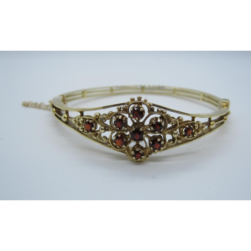 568 - A garnet set hinged Bangle, the front with openwork flower design claw-set round stones in 9ct gold.