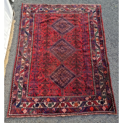 483 - A Persian style woollen red ground Rug with three central dark blue lozenges amongst multiple border... 