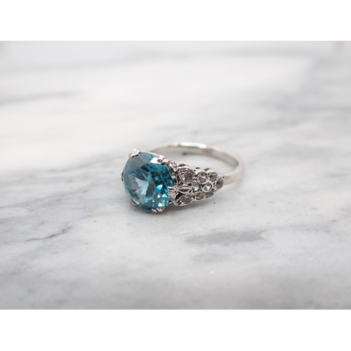 186 - A Blue Zircon Ring claw-set round stone between clusters of eight-cut white stones to shoulders in w... 