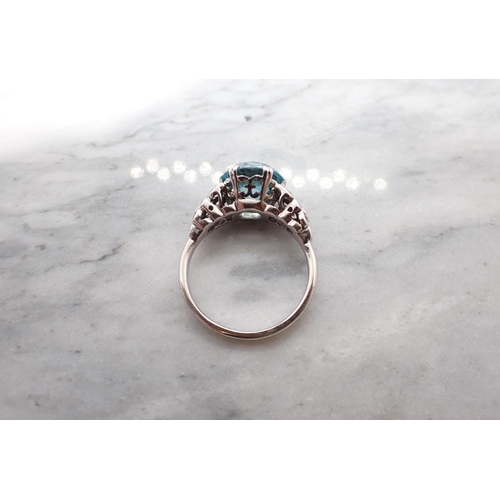 186 - A Blue Zircon Ring claw-set round stone between clusters of eight-cut white stones to shoulders in w... 