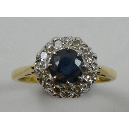 77 - An 18ct gold sapphire and diamond cluster ring, claw set with a mixed cut stone, bordered by single ... 