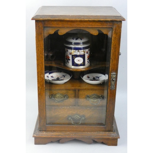 125 - An Edwardian oak smokers cabinet, the glazed door opening to reveal a tobacco jar (A/F), two ash tra... 