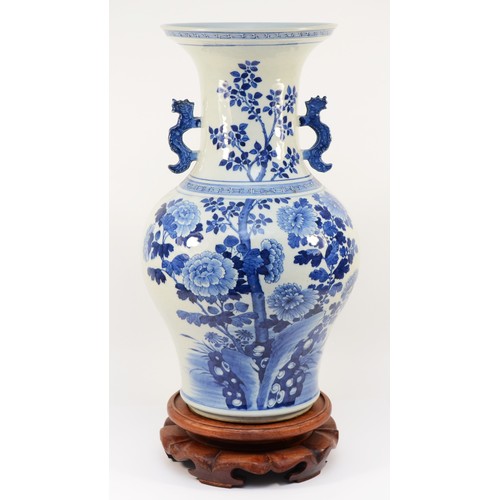 A large Chinese blue and white porcelain vase, circa 19th century Kangxi Dynasty, of baluster form, painted with a spray of lotus, peonies and prunus having Dog of Fo moulded handles, seated on a carved wood stand, export wax seals to underside, 51cm tall, hardwood stand.