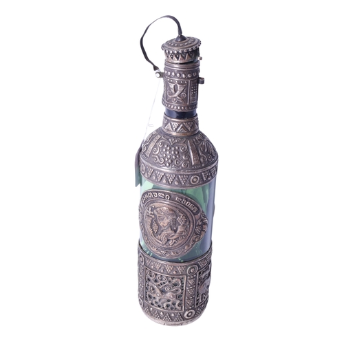 142 - A green glass bottle decorated with a white metal cover. The cover heavily decorated with embossed d... 
