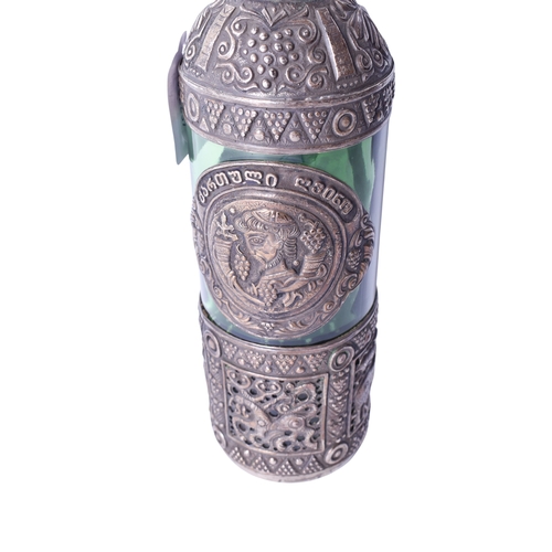 142 - A green glass bottle decorated with a white metal cover. The cover heavily decorated with embossed d... 