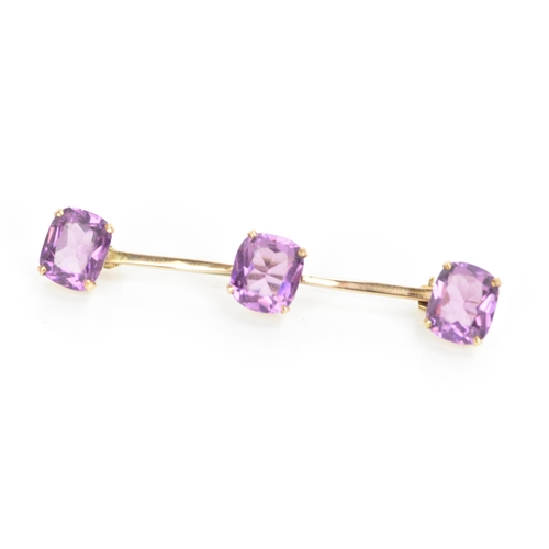 182 - An Edwardian yellow metal bar brooch, with three faceted cut amethysts in four claw settings, each s... 