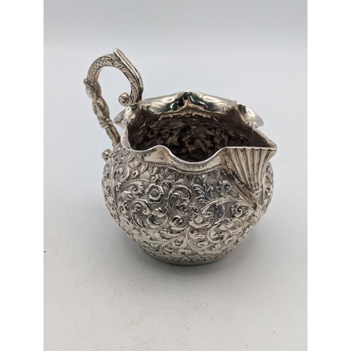 25 - An Indian white metal cream jug with deep repousse and chased continuous bands of floral scrolls, va... 