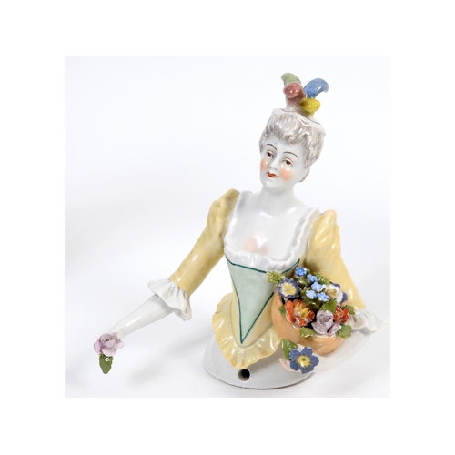 381 - A porcelain half doll, lady wearing a plumed cap and a yellow and green dress, holding a basket of f... 