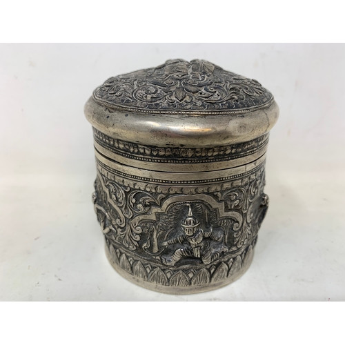 43 - A Burmese silver coloured metal cylindrical box and cover, embossed figures, the base engraved a pea... 