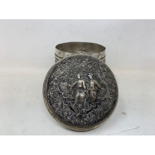 43 - A Burmese silver coloured metal cylindrical box and cover, embossed figures, the base engraved a pea... 