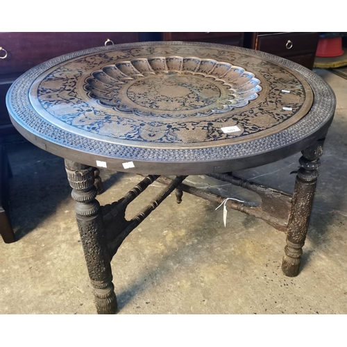 597 - Large Indian brass top and wooden framed table on ornately carved wooden base. (B.P. 21% + VAT)