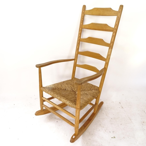 2064 - A mid-century Arts and Crafts Cotswold School style ladder-back rocking chair, in ash, with rush sea... 