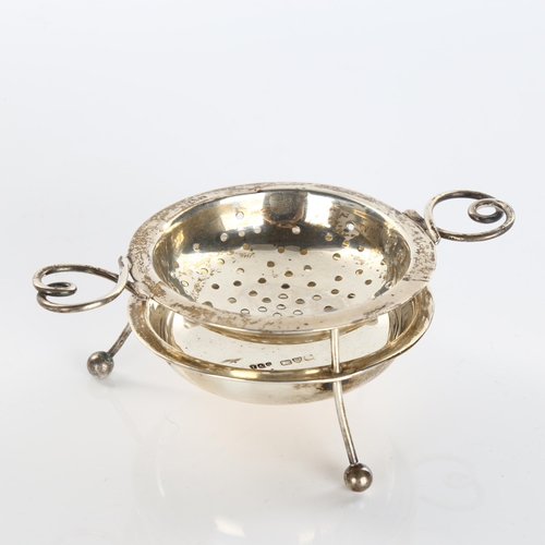 611 - An Arts and Crafts George V silver 2-handled tea strainer and stand, with wire heart-shaped handles,... 