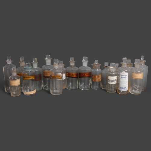 3020 - A group of 18 clear glass apothecary bottles, each with stopper, all have period labels of varying t... 