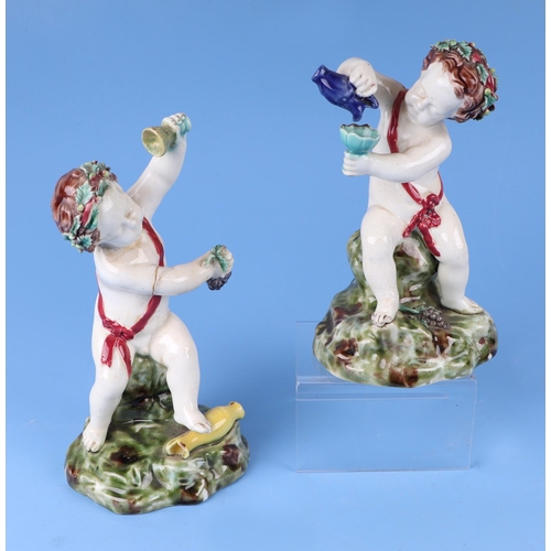 263 - A pair of continental majolica figures depicting Putti with wine glasses wearing wreaths in their ha... 