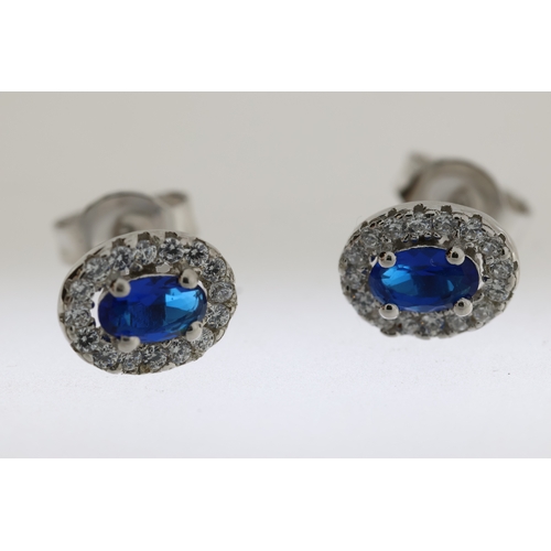 1084 - Pair of silver oval cluster studs with central blue CZ and halo of white round CZs