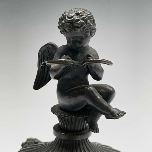 407 - A Regency bronze inkwell with cover, the lid surmounted by the figure of Cupid, the base cast with t... 