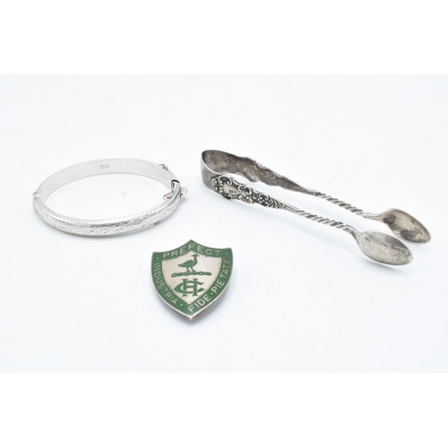 363 - A trio of silver items to include Harrogate College prefect badge, sugar tongs and a child's bangle ... 