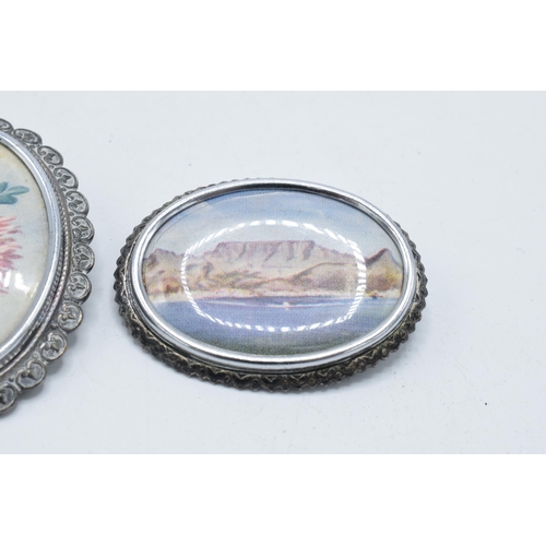 180K - A pair of brooches with a coastal and a floral scene, marked 'TLM' to rear (2).