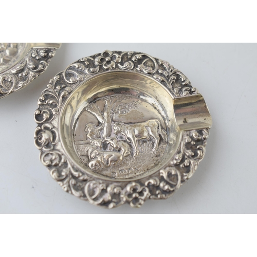 305 - A pair of sterling silver ashtrays with embossed scenes to include cows resting and a man dressed in... 