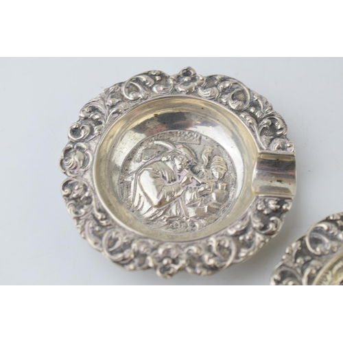 305 - A pair of sterling silver ashtrays with embossed scenes to include cows resting and a man dressed in... 