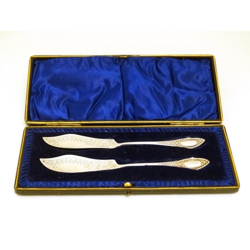 611 - A cased pair of silver plated butter knives with engraved decoration 5 3/4'' long