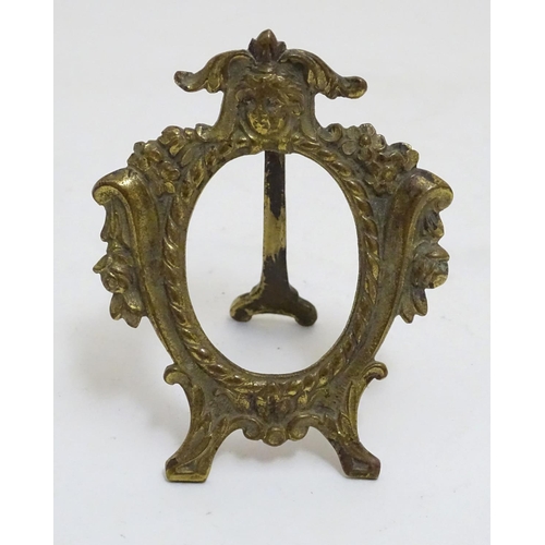 987 - A late 19th /early 20thC cast brass easel back oval frame with floral and scroll detail, surmounted ... 