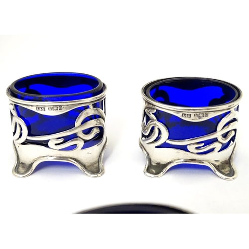 272 - An Art Nouveau silver cruet set comprising four salts and a pepper with blue glass liners and scroll... 