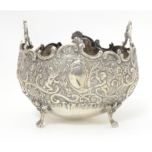 444 - A Continental white metal twin handled bowl with embossed floral, C scroll and cherub decoration, st... 