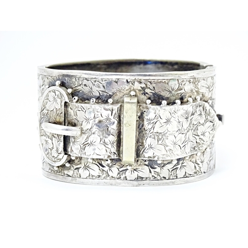 630 - A white metal bangle bracelet with buckle detail and engraved foliate decoration.