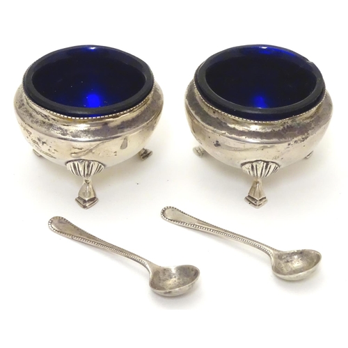 556 - A pair of circular silver table salts, each raised on three hoof feet, with blue glass liners and ma... 