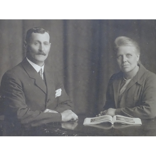 170 - An early 20thC monochrome photograph depicting a man and a woman seated at a table with an open book... 