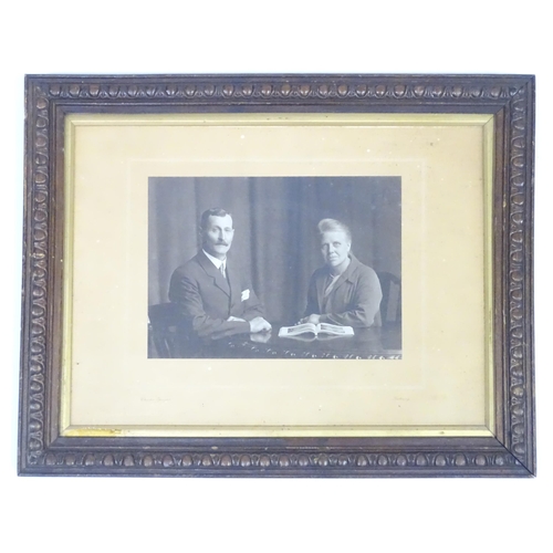 170 - An early 20thC monochrome photograph depicting a man and a woman seated at a table with an open book... 