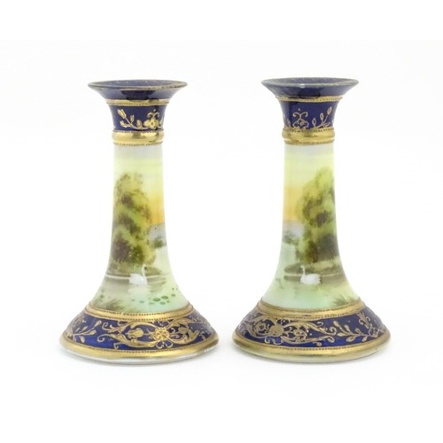 104 - A pair of Noritake style candlesticks decorated with landscape scenes with swans, bordered with gilt... 