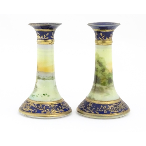 104 - A pair of Noritake style candlesticks decorated with landscape scenes with swans, bordered with gilt... 