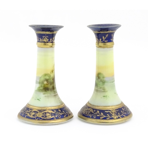 104 - A pair of Noritake style candlesticks decorated with landscape scenes with swans, bordered with gilt... 