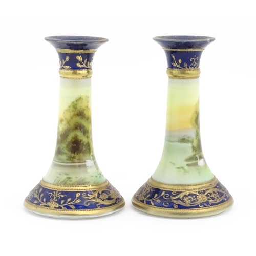 104 - A pair of Noritake style candlesticks decorated with landscape scenes with swans, bordered with gilt... 