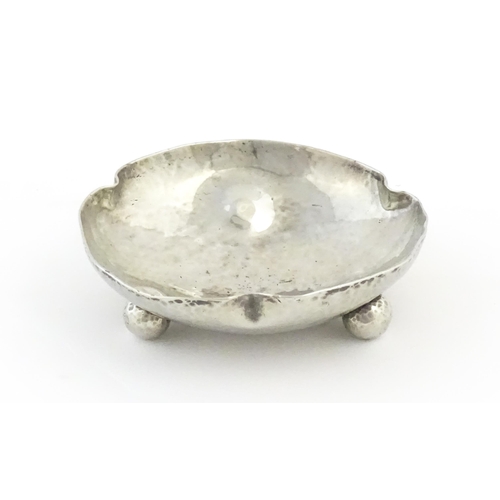 1003 - An Arts and Crafts white metal bonbon dish of shaped form with hammered decoration and three ball fe... 