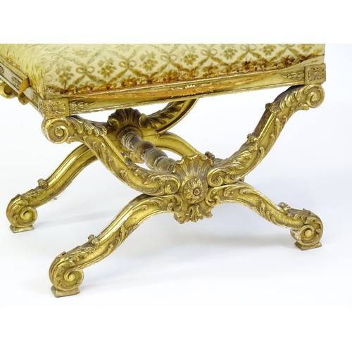 1509 - A late 19thC x-frame gilt stool, with a gesso and carved wood frame, the legs decorated with carved ... 