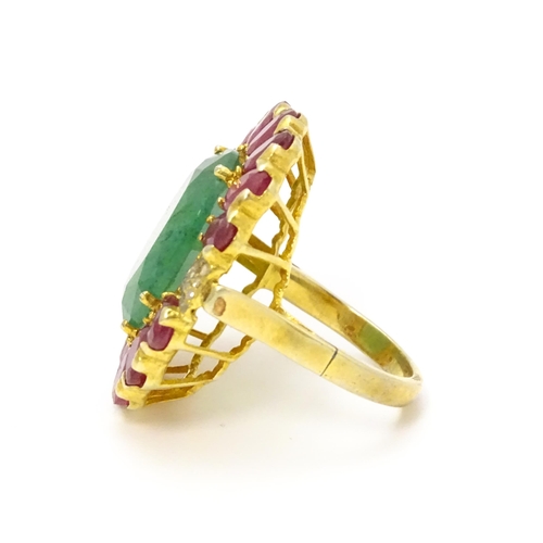 830 - A gilt metal dress ring set with central emerald coloured stone bordered by ruby coloured stones fla... 