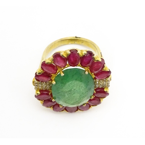 830 - A gilt metal dress ring set with central emerald coloured stone bordered by ruby coloured stones fla... 