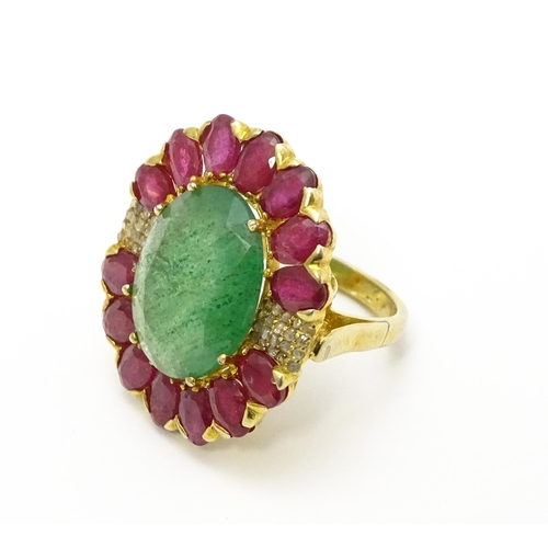 830 - A gilt metal dress ring set with central emerald coloured stone bordered by ruby coloured stones fla... 