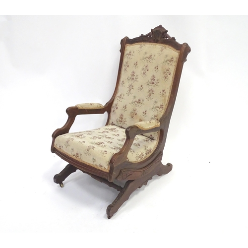 643 - An early 20thC rocking chair with carved cresting rail above a chamfered frame, with upholstered bac... 