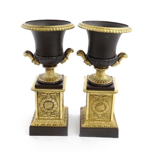 1019 - A pair of cast bronze campana urns with twin handles raised on squared plinths with relief symbols o... 